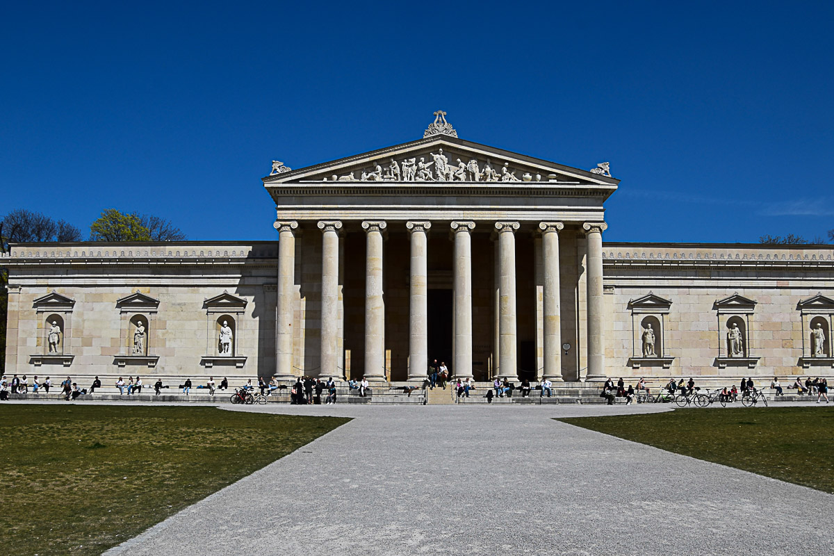 Blick auf die Glyptothek am Königsplatz, ein Bau im Stil der griechischen Antike mit Säulen und Skulpturengiebel. Menschen genießen die Sonne auf den Treppen.